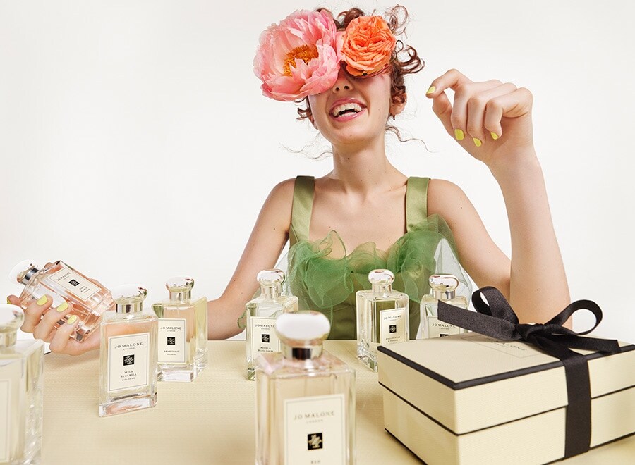 Mulher sorrindo com flores no rosto e segurando uma colônia e várias colônias de Jo Malone na frente com uma caixa de presente