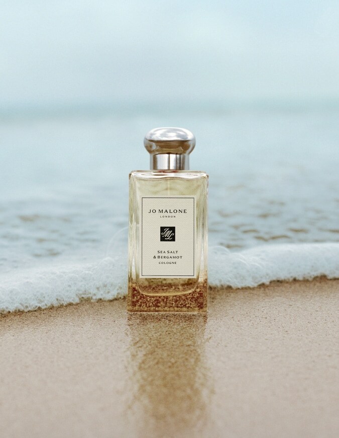 Jo Malone London Sea Salt & Bergamot cologne standing on wet sand with gentle waves behind.