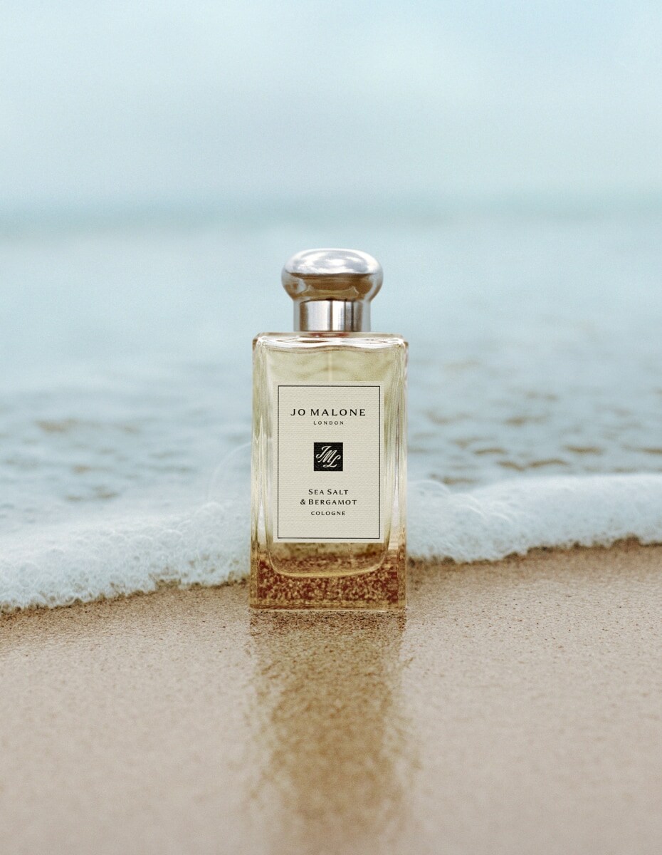 Jo Malone London Sea Salt & Bergamot cologne standing on wet sand with gentle waves behind.
