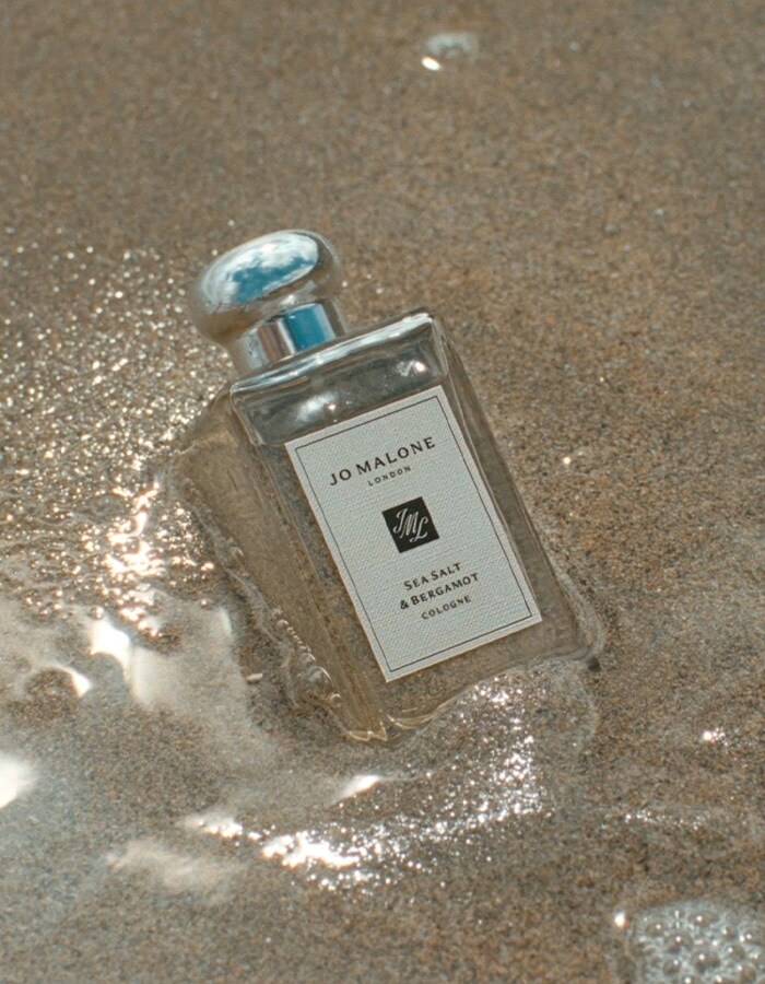 Jo Malone London Sea Salt & Bergamot cologne standing on wet sand with gentle waves behind.