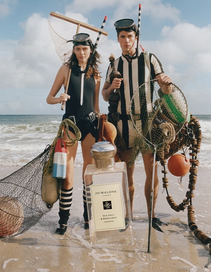 Two people in swimwear hold nets and buoys on beach with oversized Jo Malone London cologne bottle.