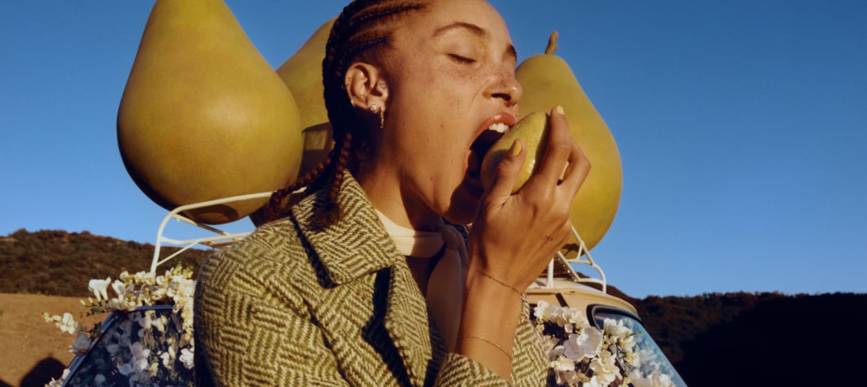 Adwoa Aboah with braided hair, biting into a pear with backdrop of three large pear props placed on top of a car roof rack.